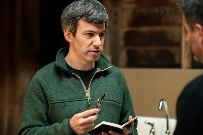 Nathan Fielder in a green zip-up fleece with a pencil and notebook, addressing another person.