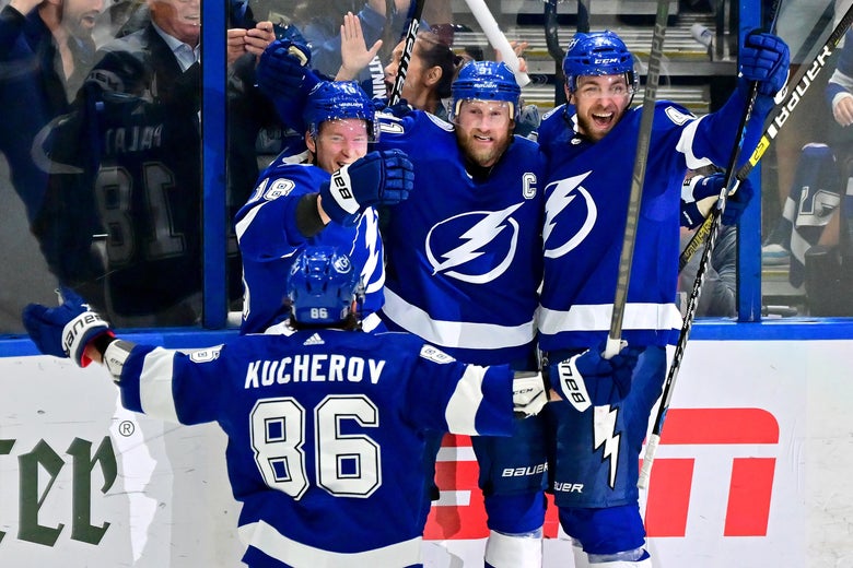 Stamkos, Rutta, and Palat smile and hold their arms and sticks up in celebration, Kucherov seen from behind approaching them with his arms extended out wide to his sides