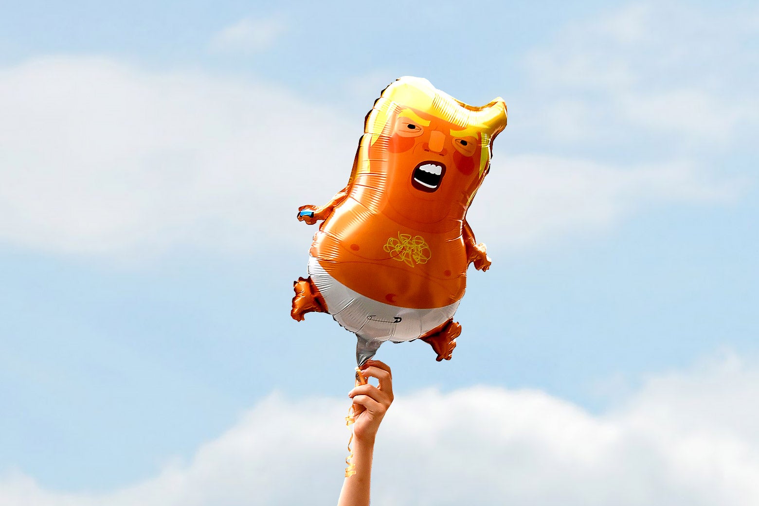A hand holds up a balloon against a blue sky with clouds. The balloon is an image of Donald Trump as a baby, yelling, naked except for a diaper.