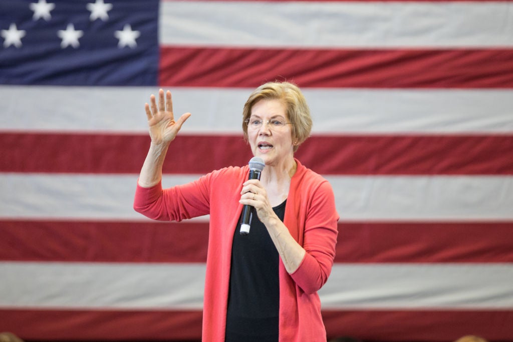 Warren speaks into a mic onstage, with an American flag behind her.