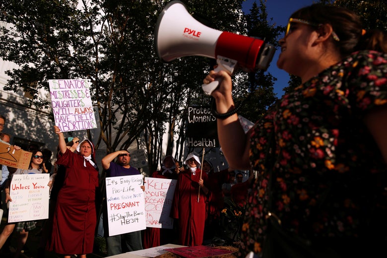 Protesters carry pro-choice signs; some are wearing red Handmaid's Tale dresses. In the foreground, a protester shouts into a megaphone.