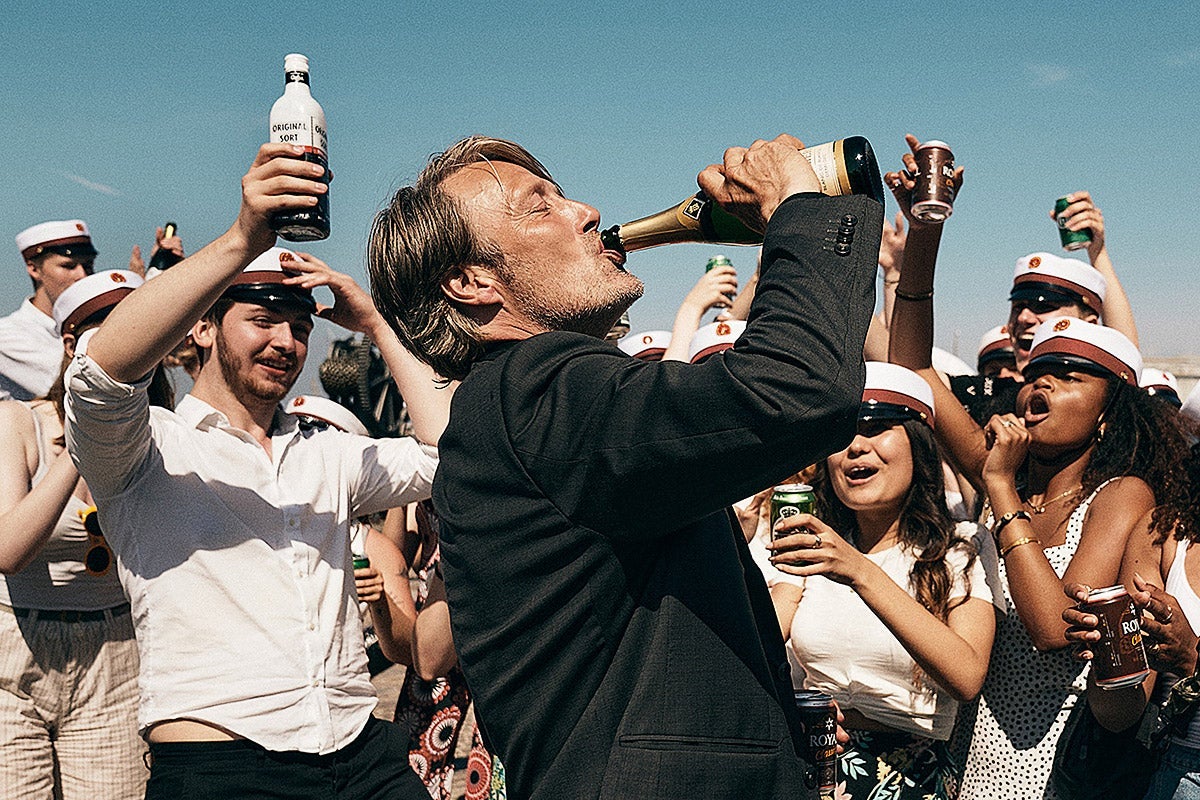 Mads Mikkelsen drinks from a bottle as a crowd cheers him on.