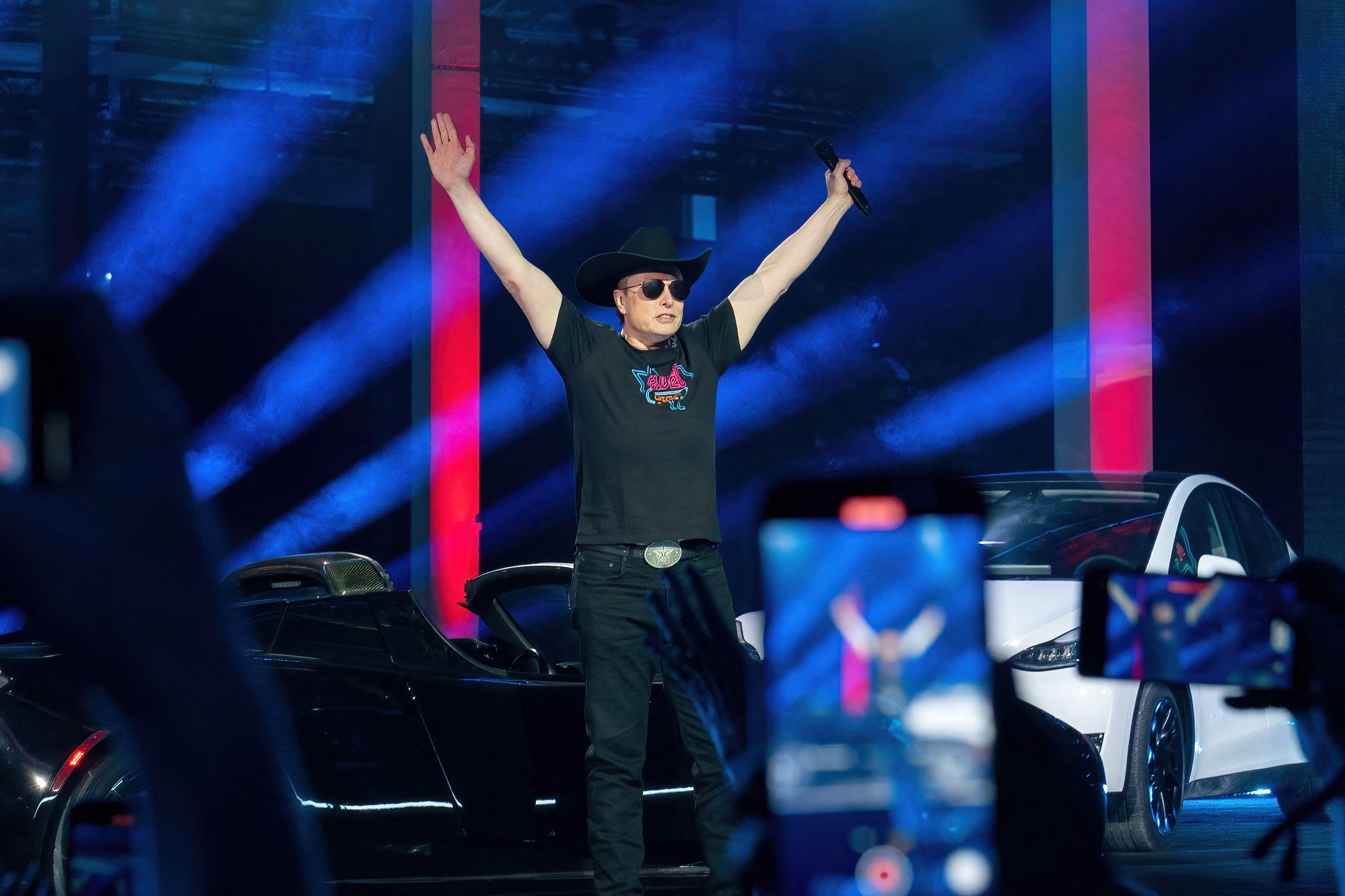 Elon Musk, wearing a cowboy hat, raises his arms up wide while standing on a stage in front of cars.