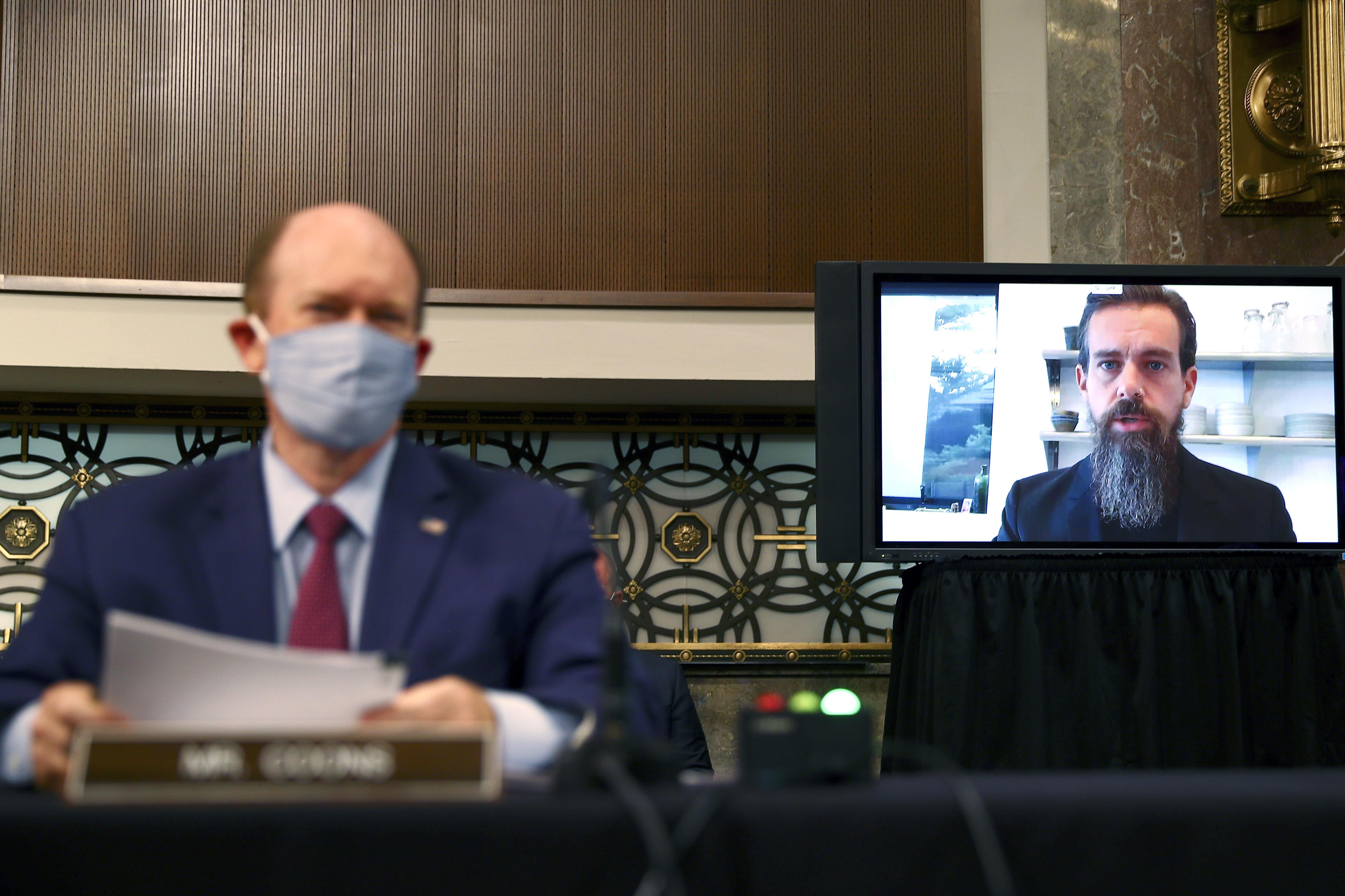 On the left, an out-of-focus man sitting behind a nameplate; on the right, a man with a beard appears on a TV screen.