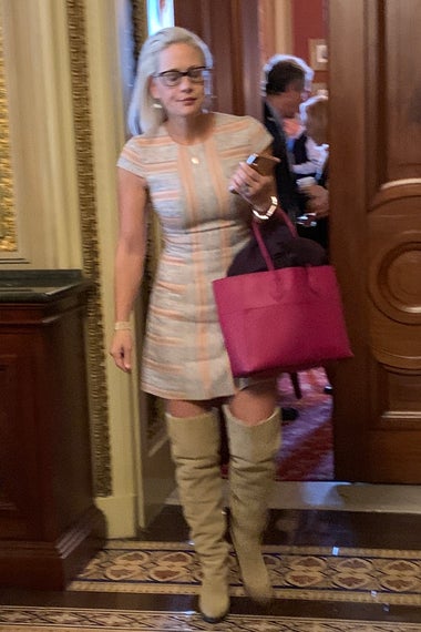 Kyrsten Sinema in a pastel striped minidress, a hot pink tote, and taupe over-the-knee boots.