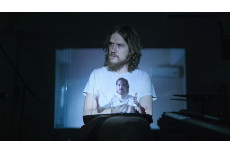 An unkempt Bo Burnham sits in a chair wearing a white t-shirt, on which is projected an image of Bo Burnham giving a double thumbs-up.