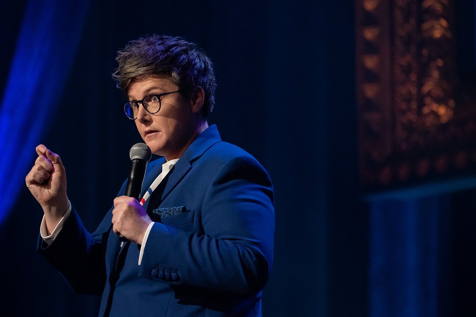 Hannah Gadsby onstage wearing a blue jacket and striped shirt, holding a mic.