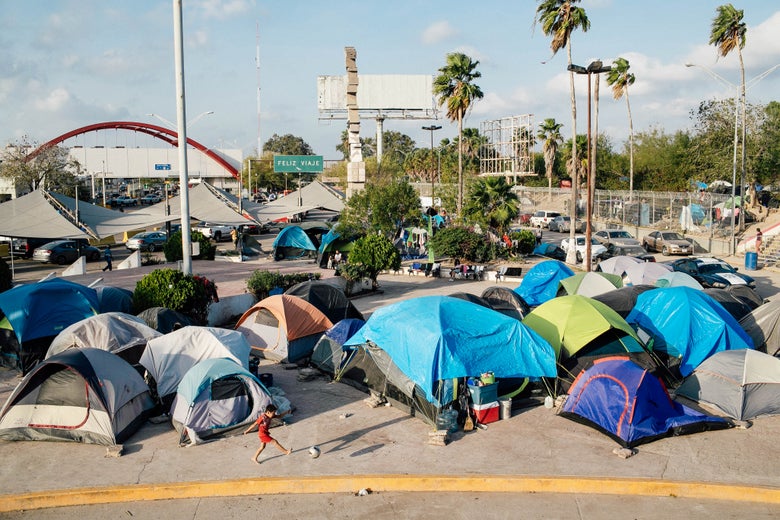 A boy kicks a soccer ball in front of a grouping of tents in front of a bridge.