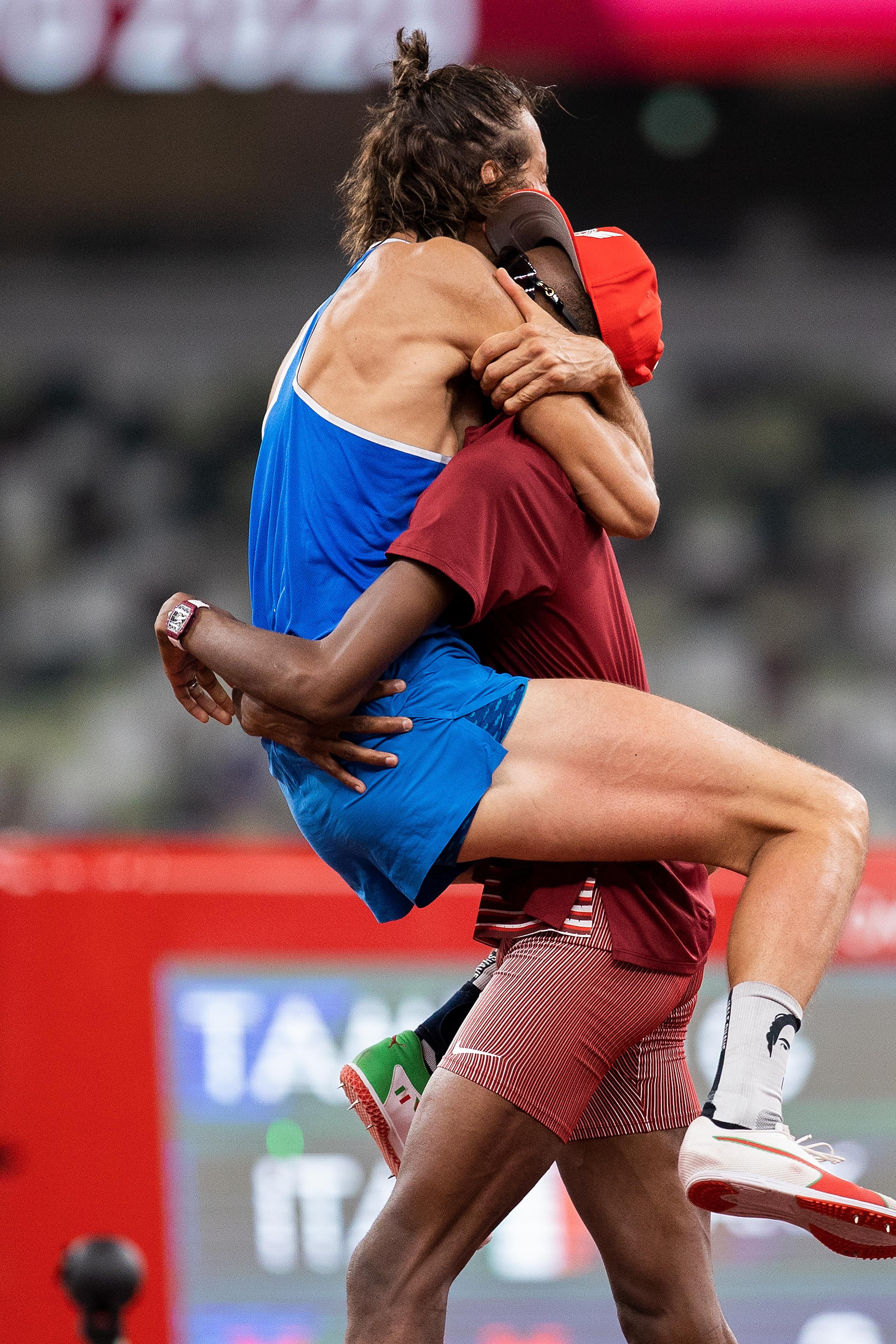 A high jumper wearing a blue uniform leaps into the arms of a high jumper wearing a red uniform. They embrace.