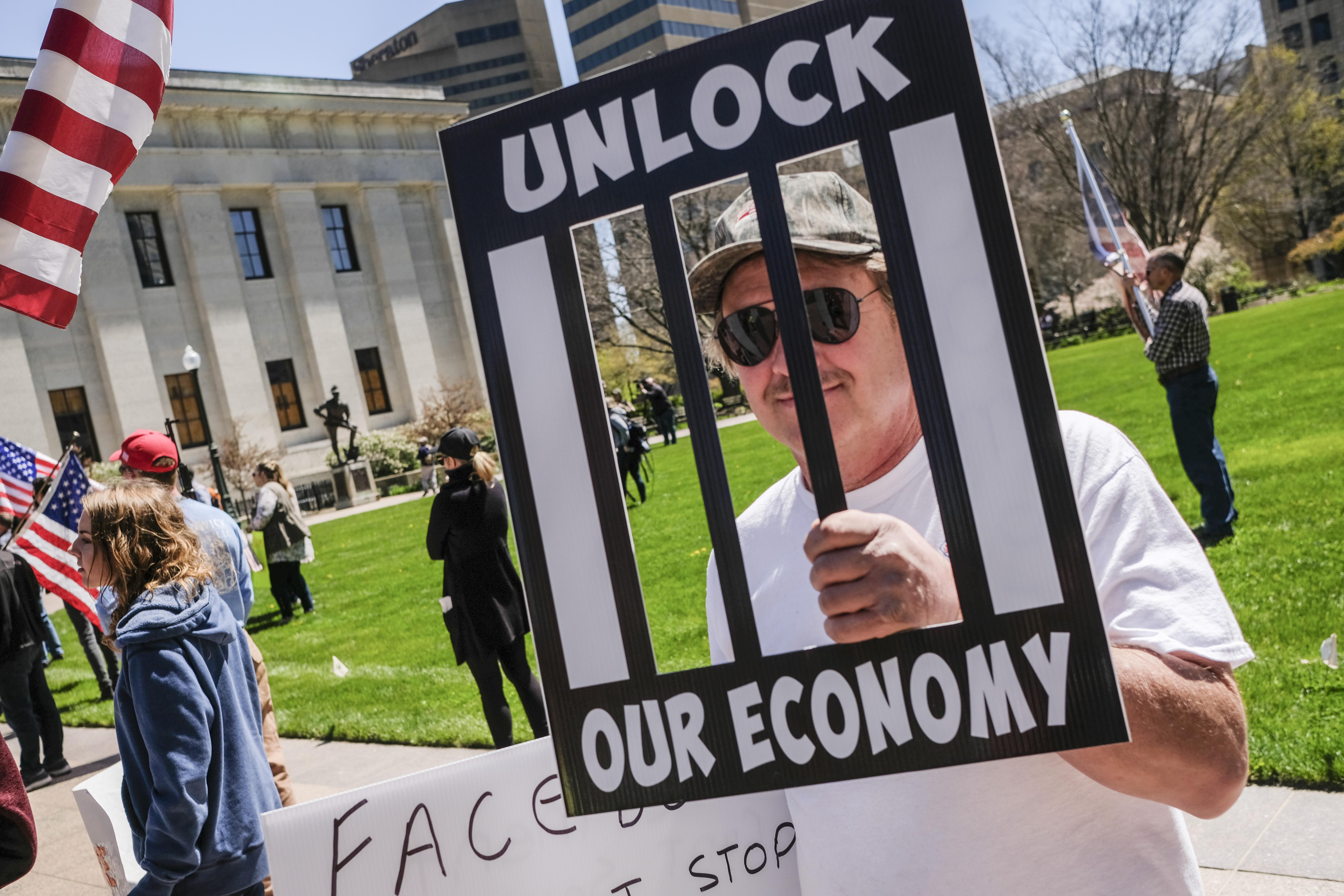 A man in a white t-shirt and sunglasses carries a sign that reads "UNLOCK OUR ECONOMY."
