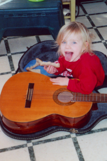 A young, blonde-haired girl sticks her tongue out at the camera, holding a sandwich crust and leaning on an adult-size guitar.
