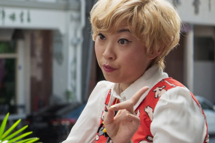Awkwafina wears a short blonde wig and holds up a finger to someone off-camera.