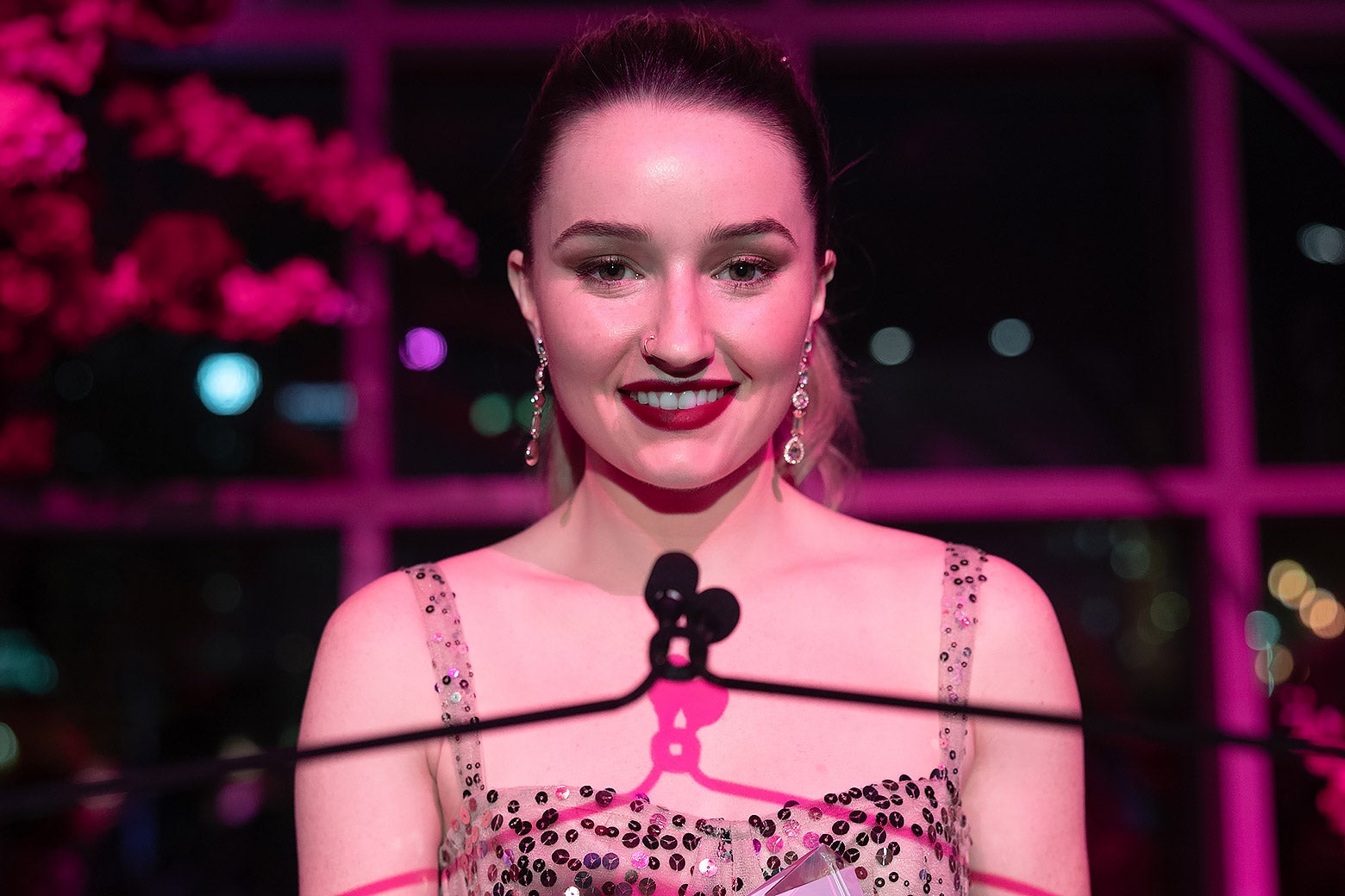 Kaitlyn Dever as Belle Gibson, standing at a podium under a purple-tinted light.