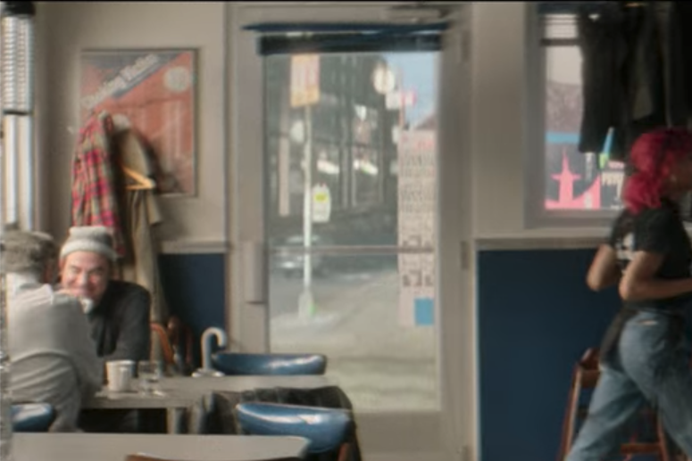 James C. Nicola with his back facing the camera dines at a diner table with Roger Bart who is facing the camera. 