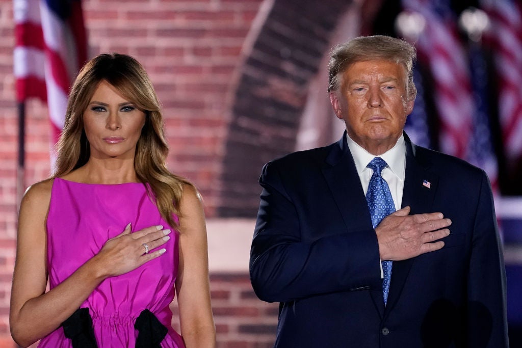 The Trumps, lit by stage lights, stand side by side in front of a brick arch holding their hands over their hearts.