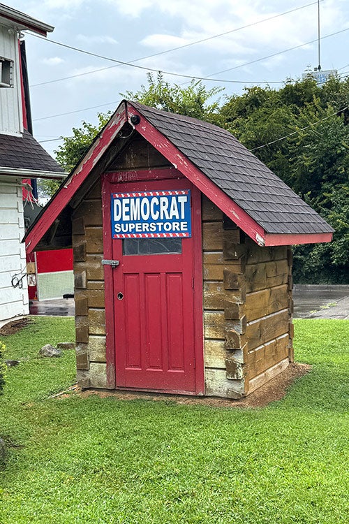 A tiny wooden log cabin on a grassy lawn. Mounted on the door is a blue-and-white sign that reads "DEMOCRAT SUPERSTORE."
