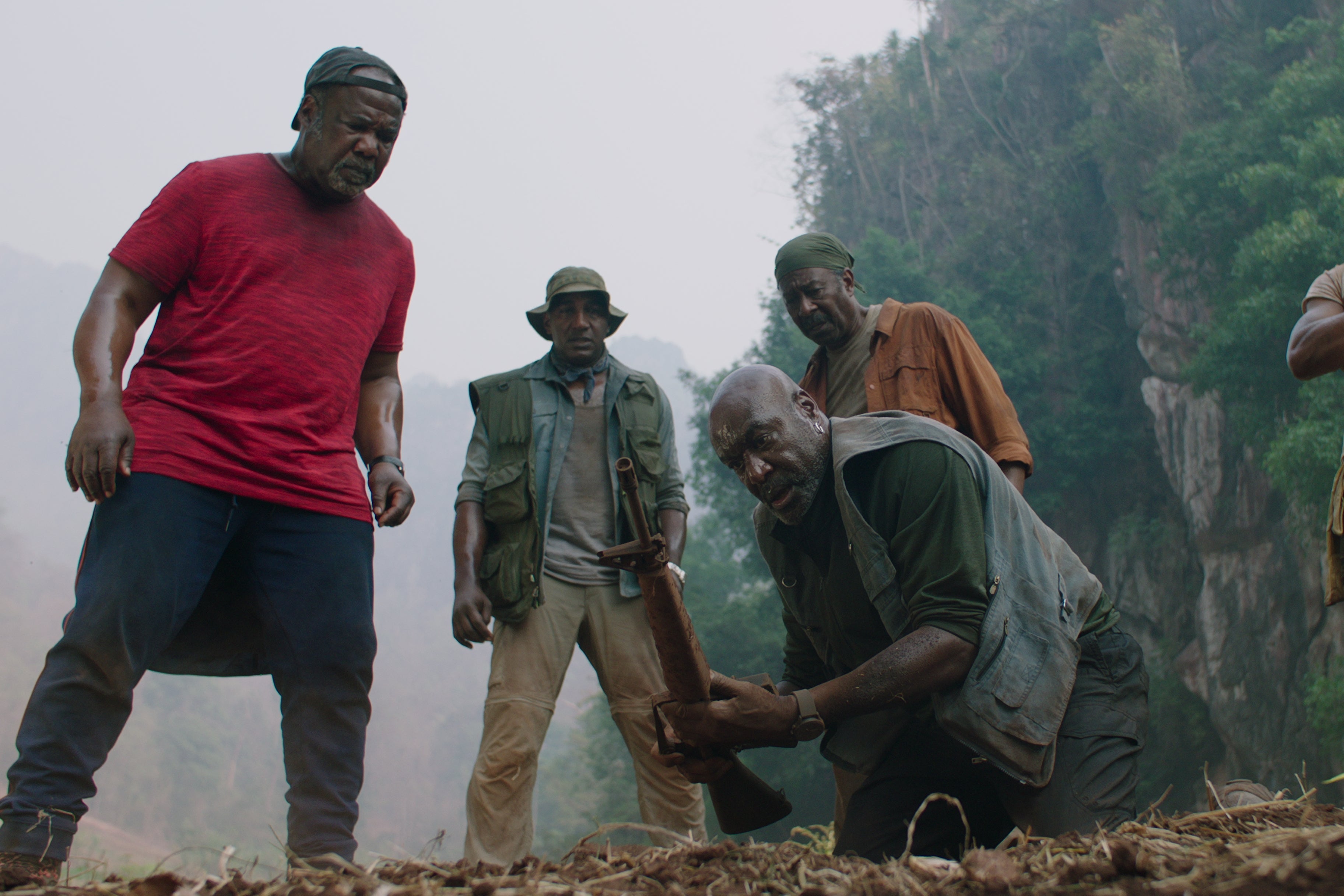 Men gather around Delroy Lindo as he lifts a rifle from the ground