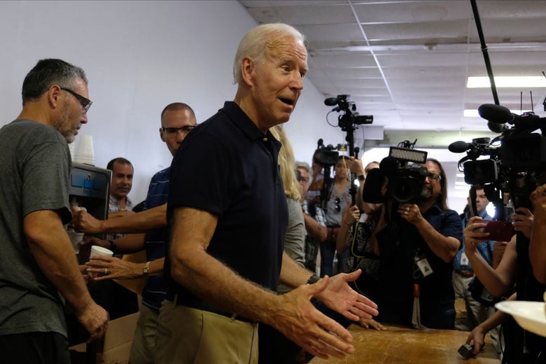 Biden, wearing a blue polo shirt and khakis, gestures while speaking in front of reporters and camera operators.