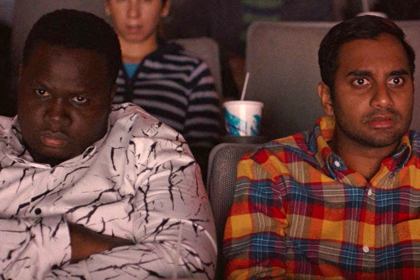 A Black man and an Indian American man sit in a movie theater and look ahead in dismay.