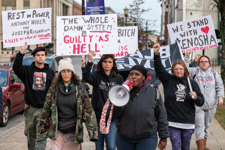 A group of people hold up signs. A Black woman at the front speaks into a megaphone.