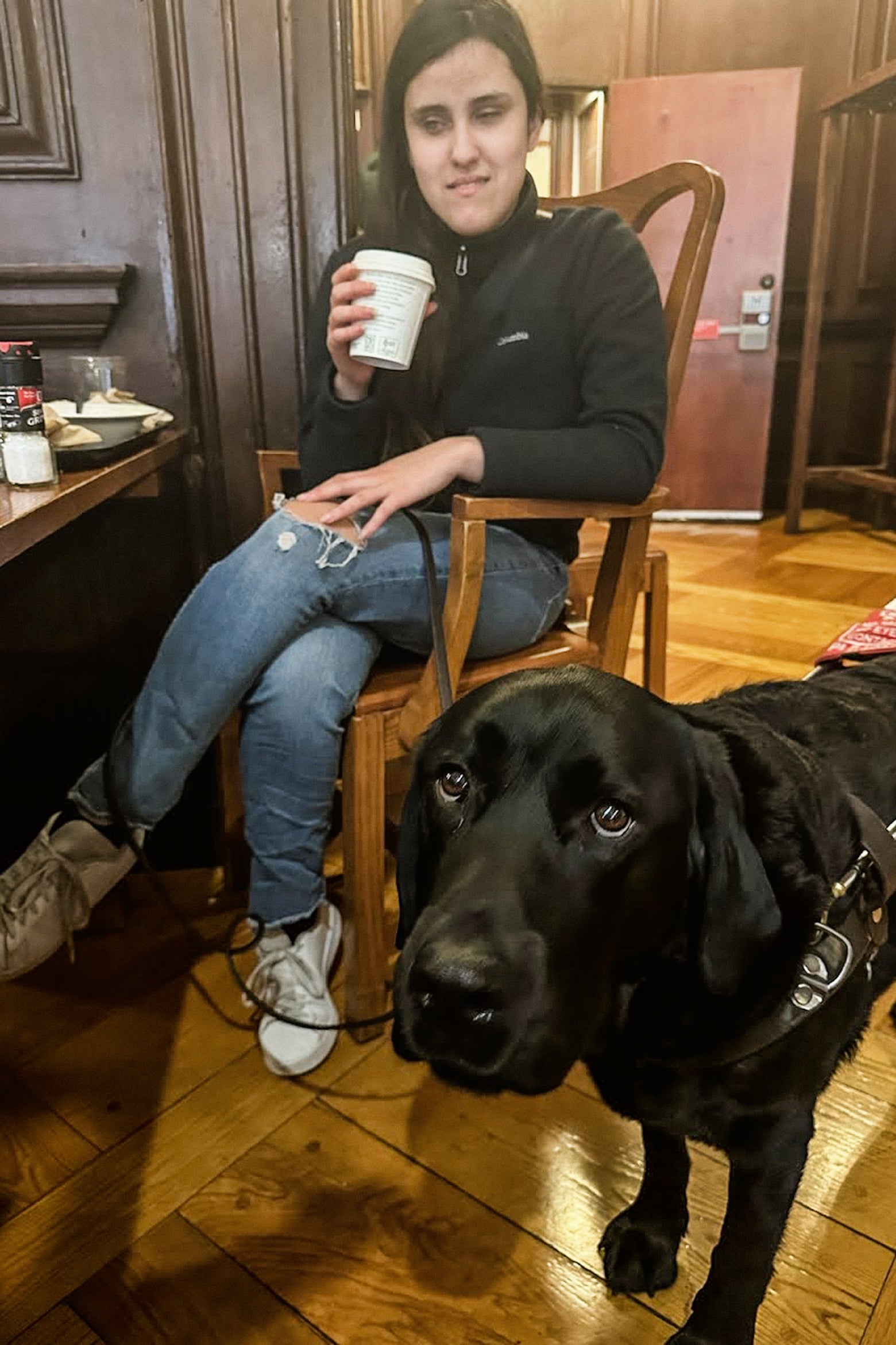The picture shows a young woman sitting on a wooden chair in what appears to be a cozy café with wooden interiors. She is wearing a black jacket and blue jeans, and holding a white paper cup, possibly with a hot beverage. She has a somewhat serious expression on her face. To her right, there is a black Labrador dog standing on the wooden floor, looking directly at the camera. The dog is wearing a guide dog harness and looks serious. On the table next to the woman, there are condiments like salt and pepper, and a red bag of chips. The atmosphere seems relaxed and casual.