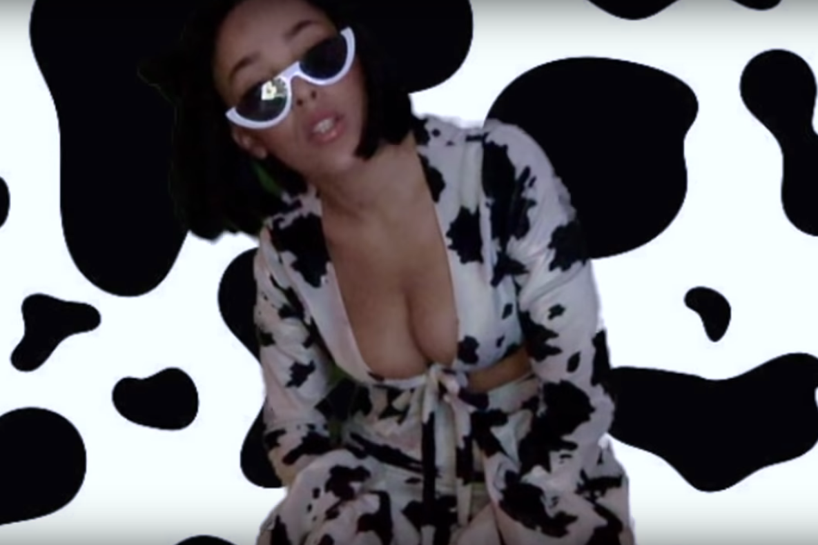 Doja Cat stands in a cow costume in front of a background imitating a cow's spots.
