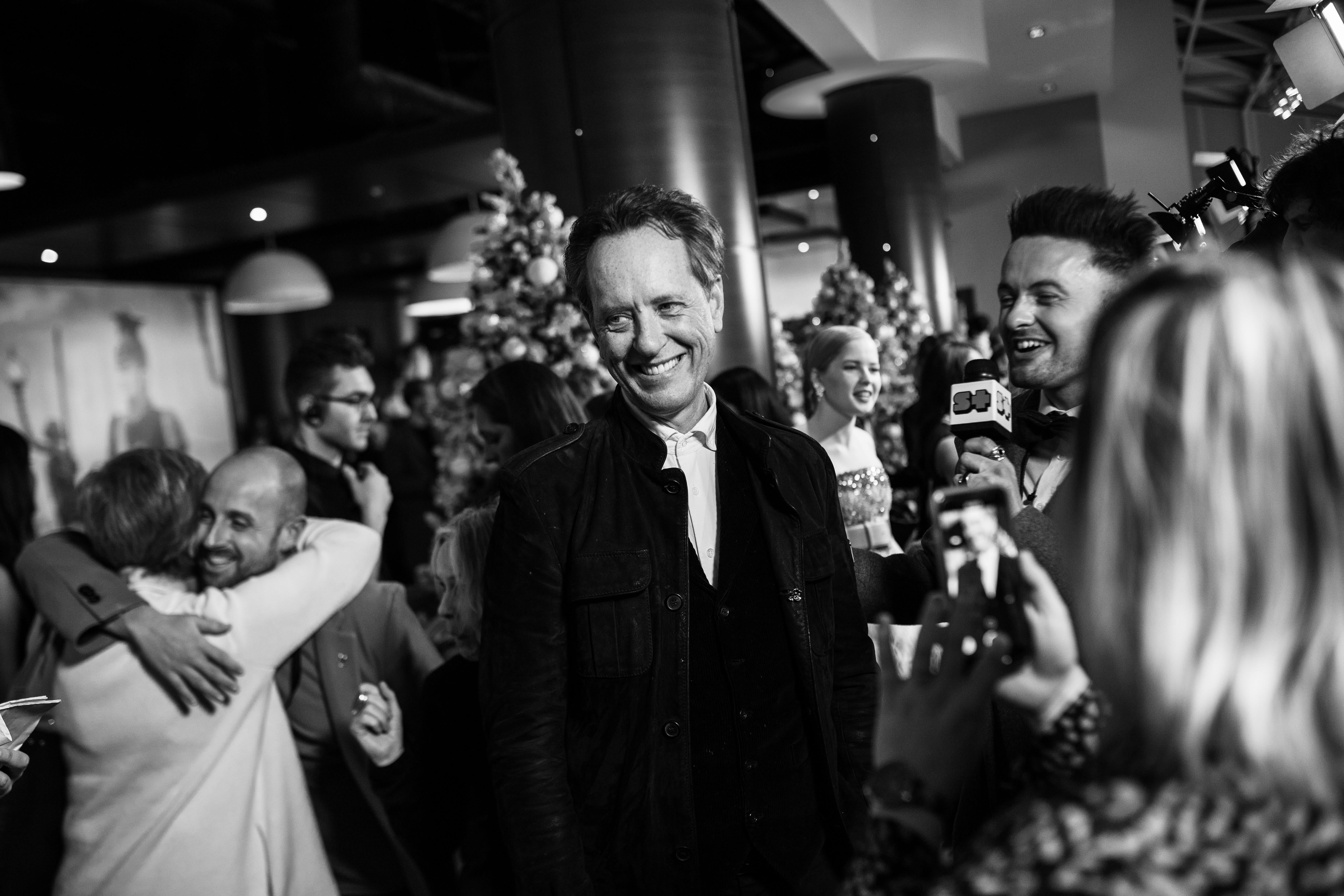 A black-and-white photo of Richard E. Grant smiling, surrounded by other people on a crowded red carpet for a movie premiere in November 2018.