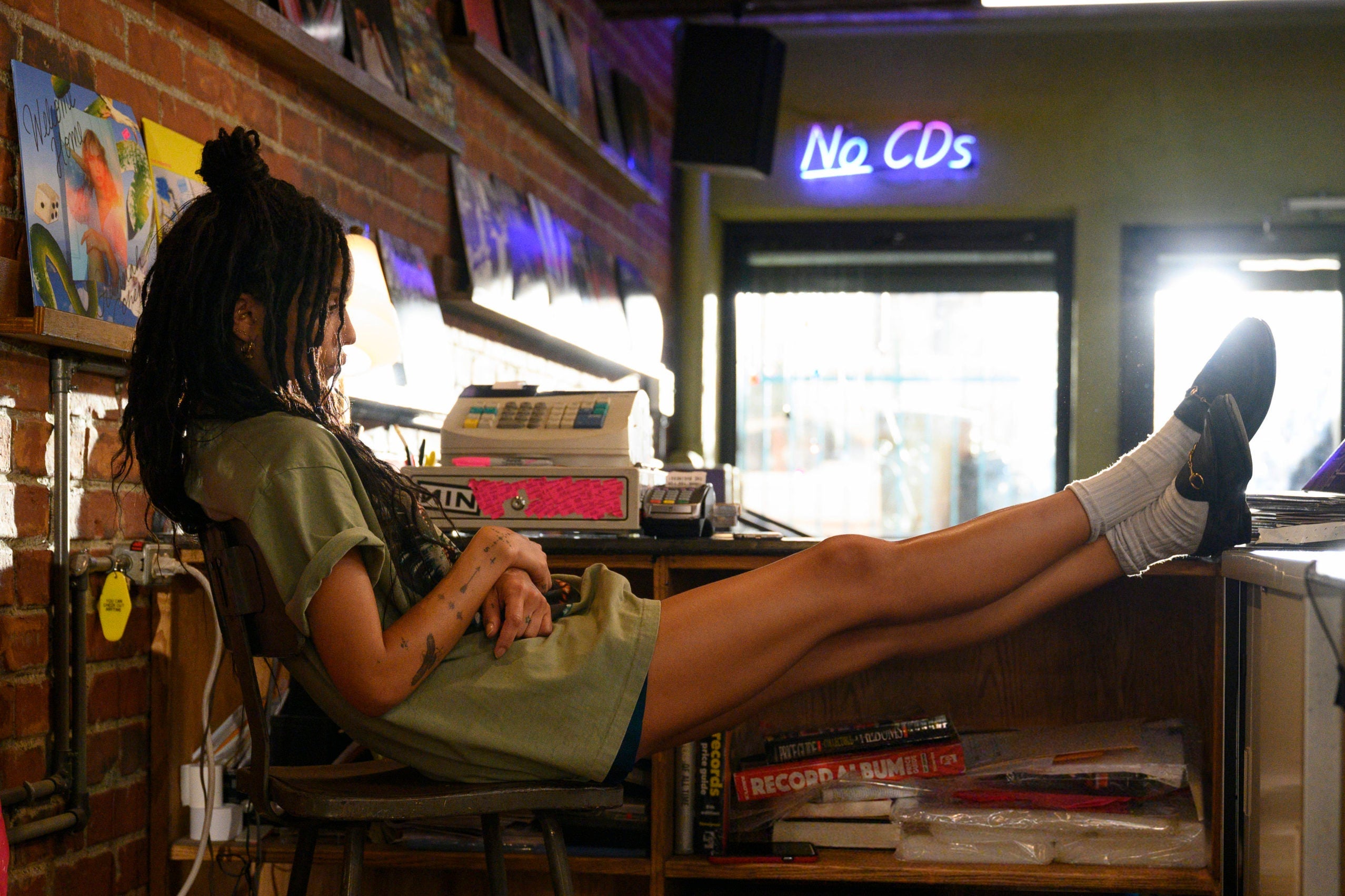 Zoë Kravitz in a scene from High Fidelity, in which she sits at a cash register while a "No CDs" neon sign blares in the background.