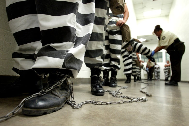Incarcerated people in striped uniforms stand in a line with their legs chained in a hallway