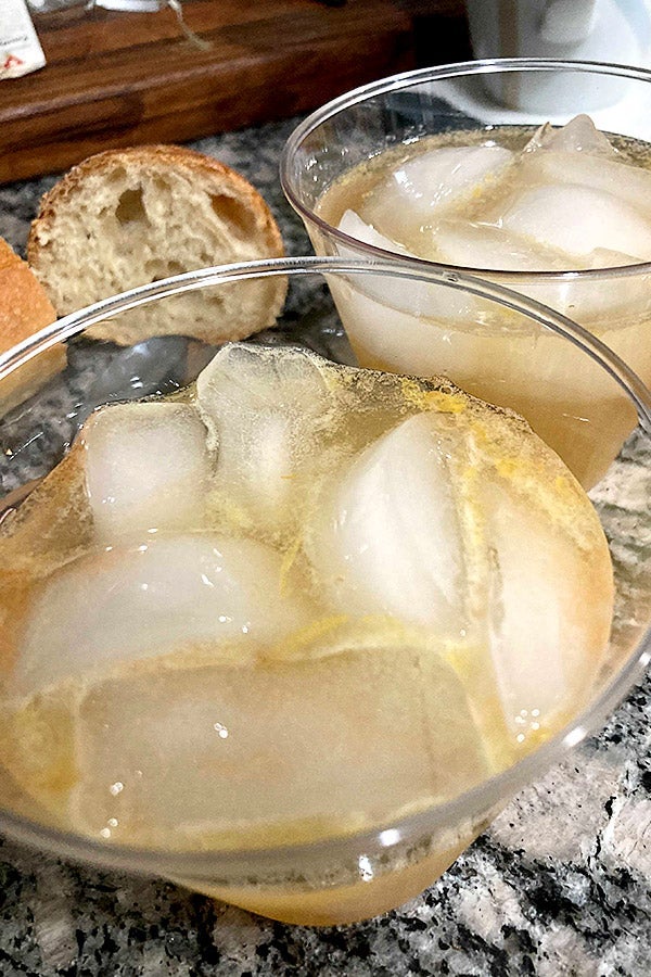 Two glasses of a brown liquor with numerous ice cubes in them sit on a counter. There is a piece of bread to the side of them. 
