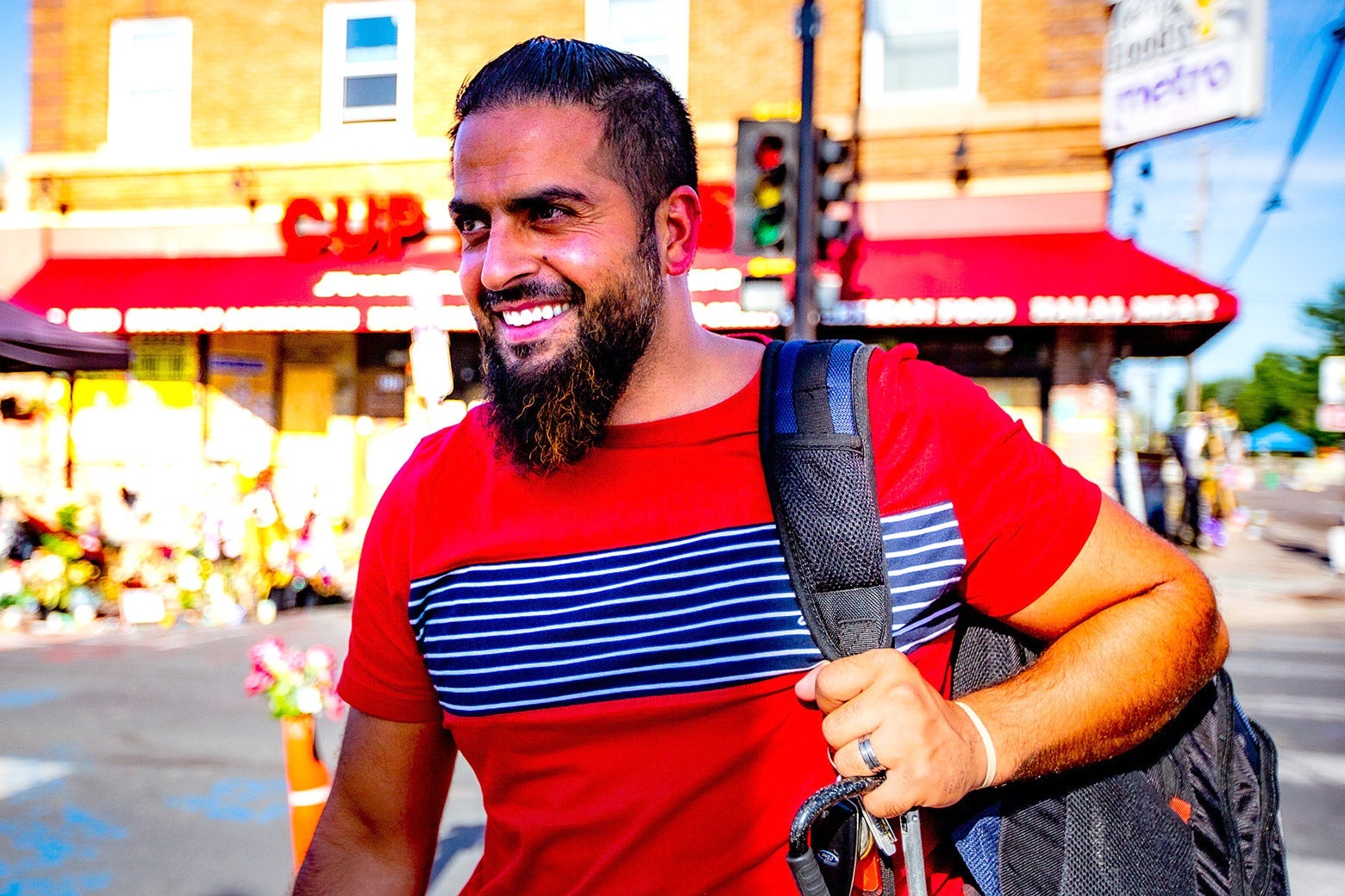 Mahmoud Abumayyaleh in a red T-shirt with a backpack over one shoulder. The CUP Foods store is behind him.