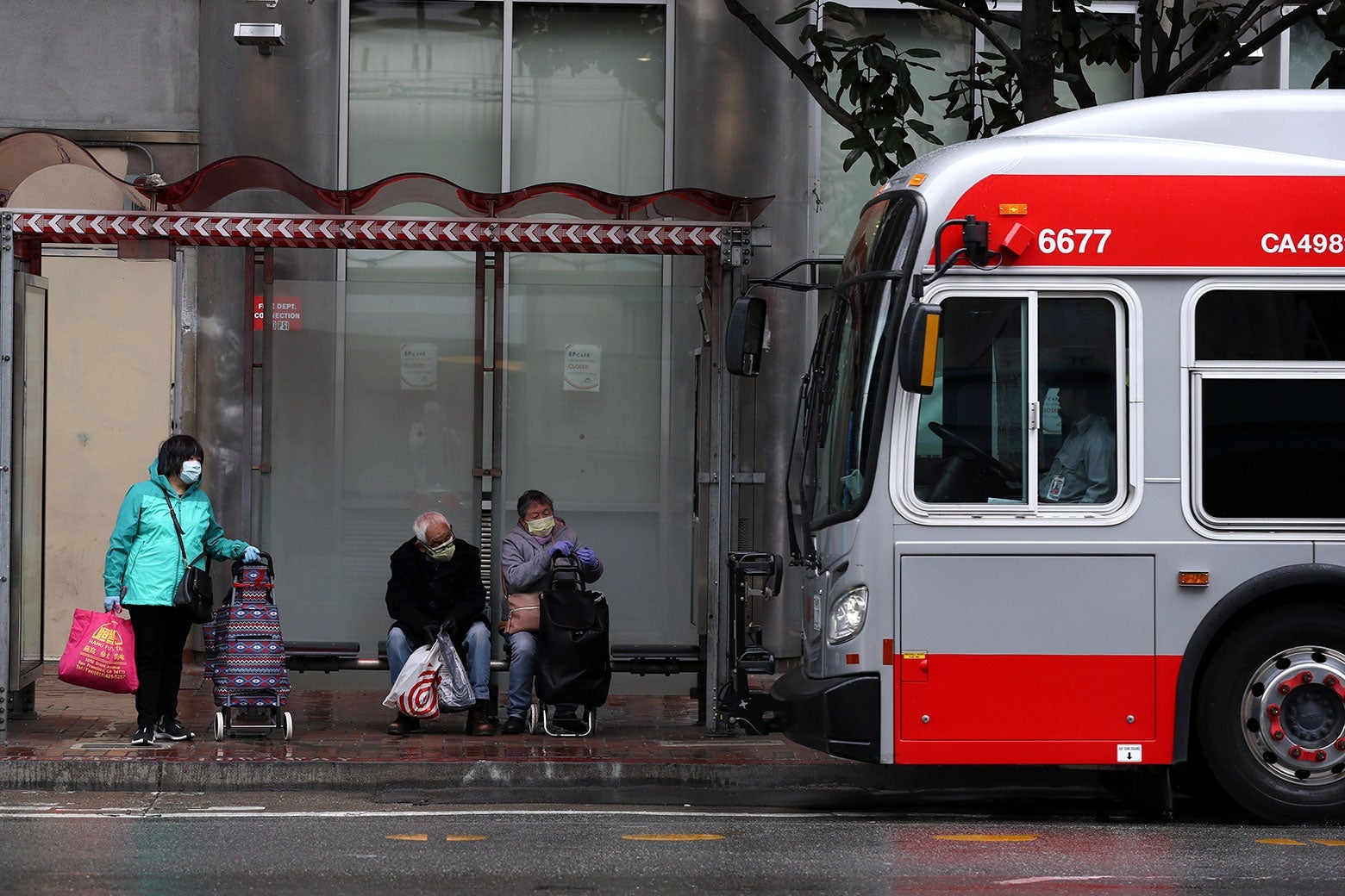Riders, in face masks, wait for a MUNI bus as it pulls into a stop in San Francisco. Looks like a gray day.