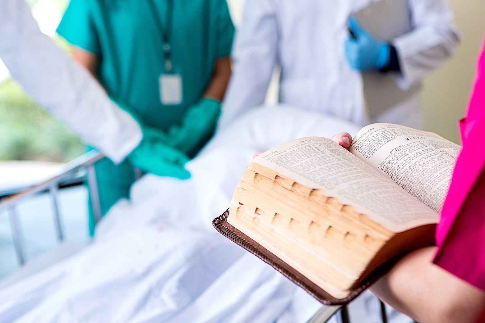 Doctors stand at the foot of a bed and put their hands on a patient's body. Across from the, a person holds open a heavy book.