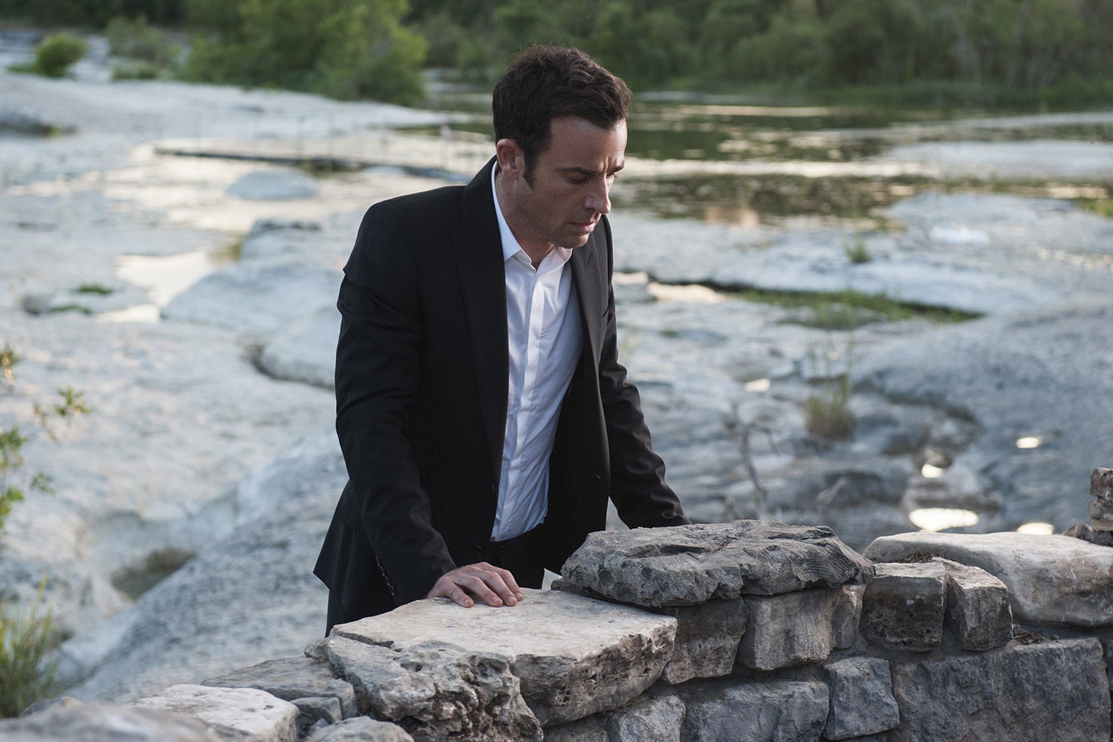 A white man in a suit stands on a bridge and stares in confusion below.