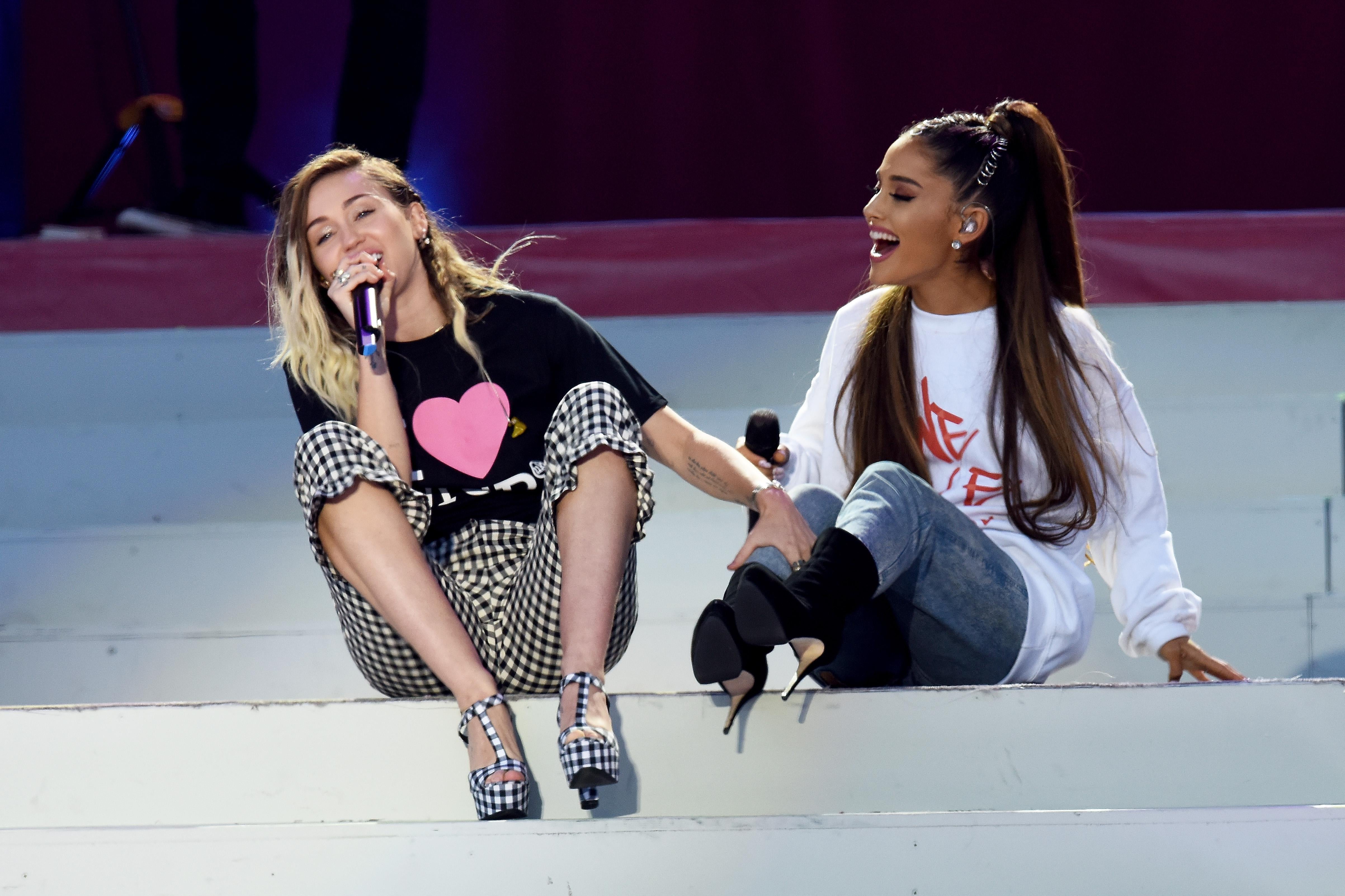 Miley Cyrus and Ariana Grande smile and sit on the white stairs while performing onstage together