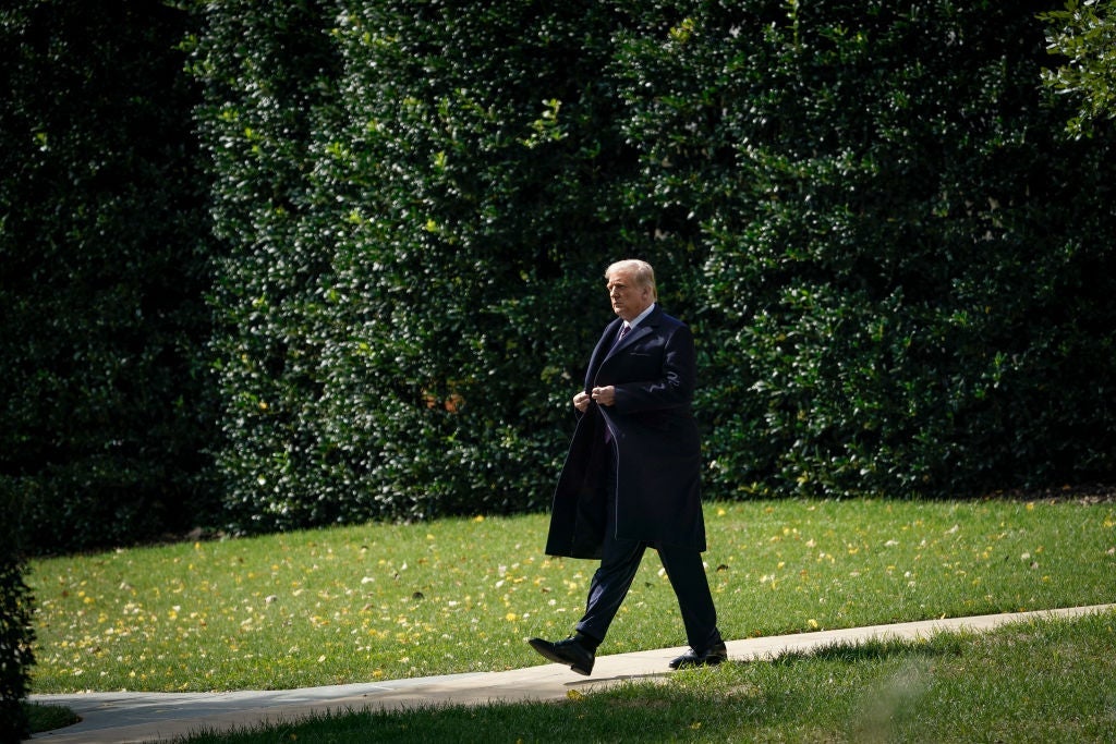 Trump, wearing a suit and a dark coat, walks by himself across a sidewalk against a backdrop of foliage.