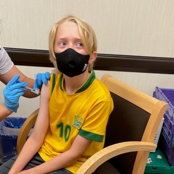 A boy wearing a yellow soccer jersey sits in a chair and receives a vaccine.