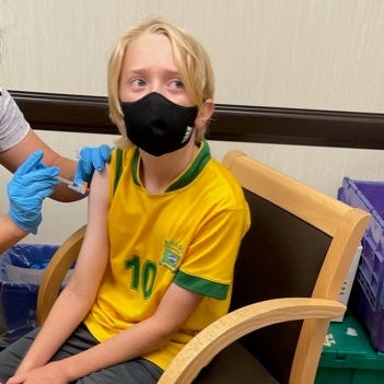 A boy wearing a yellow soccer jersey sits in a chair and receives a vaccine.