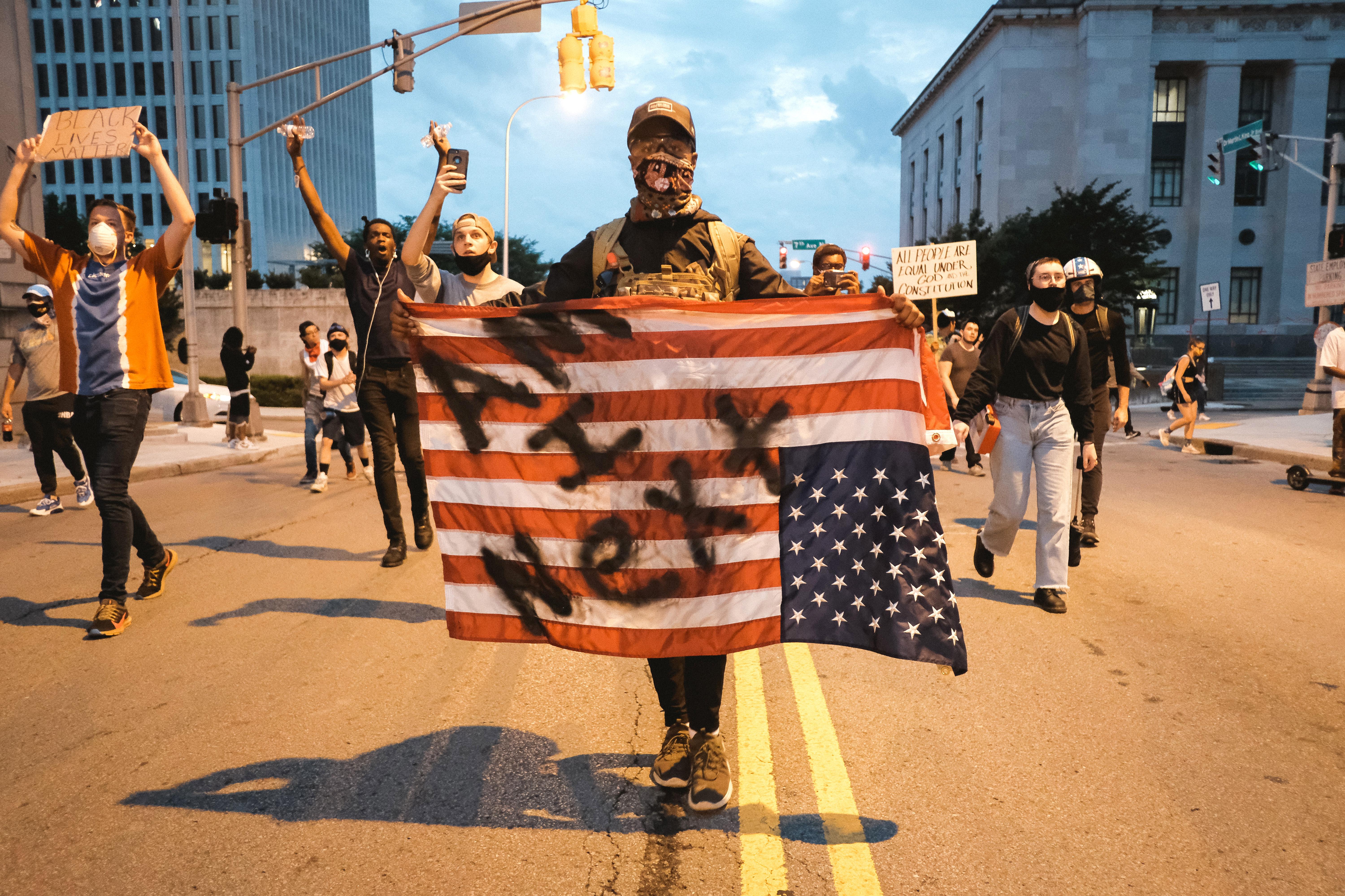 A man in a bandana holds an upside-down U.S. flag on which can be seen the words "Am I Next?" Other protesters walk on the street and hold signs behind him.