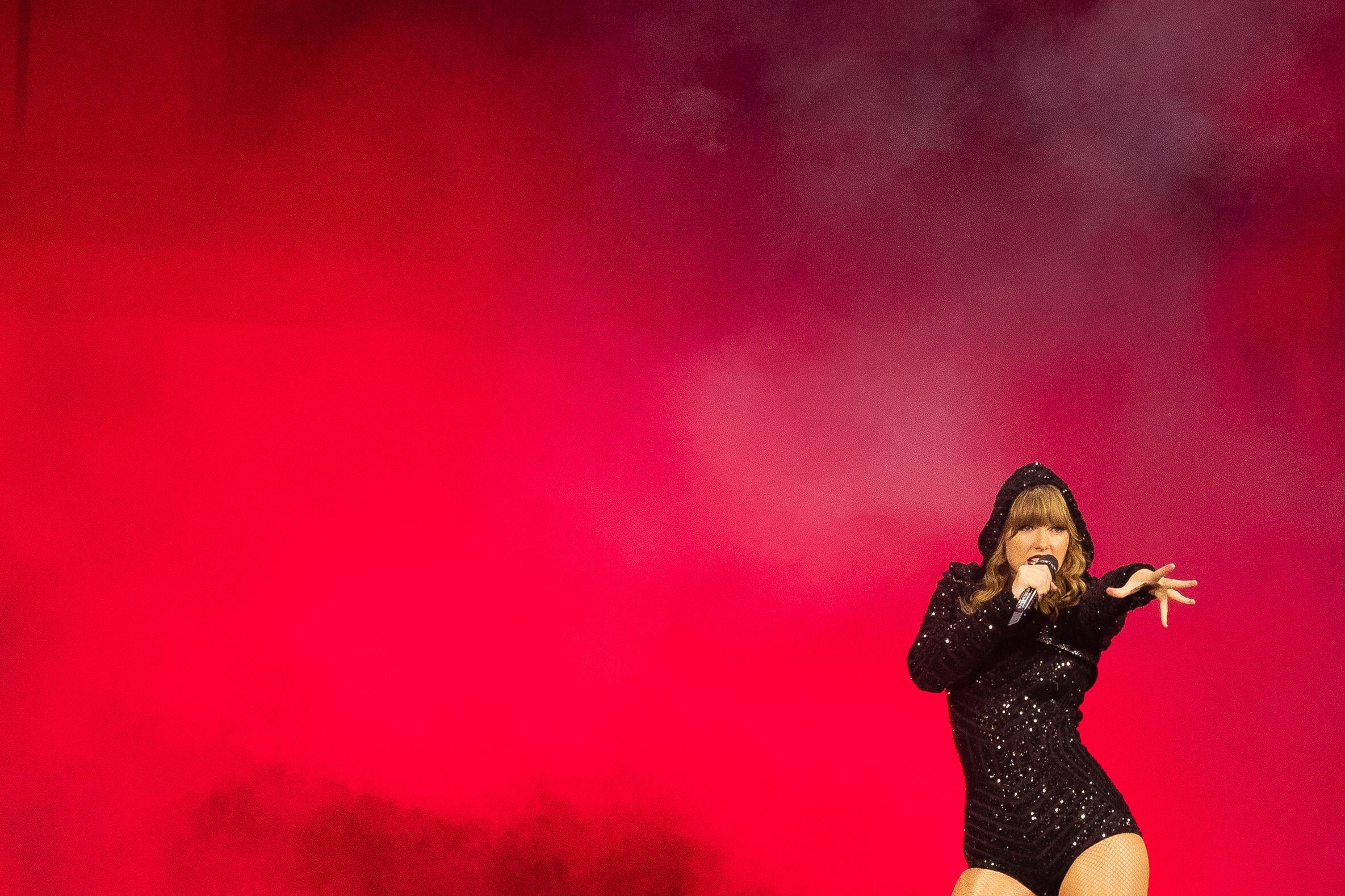 Taylor Swift, wearing a glittery, hooded leotard, stands in front of a wall of red smoke.