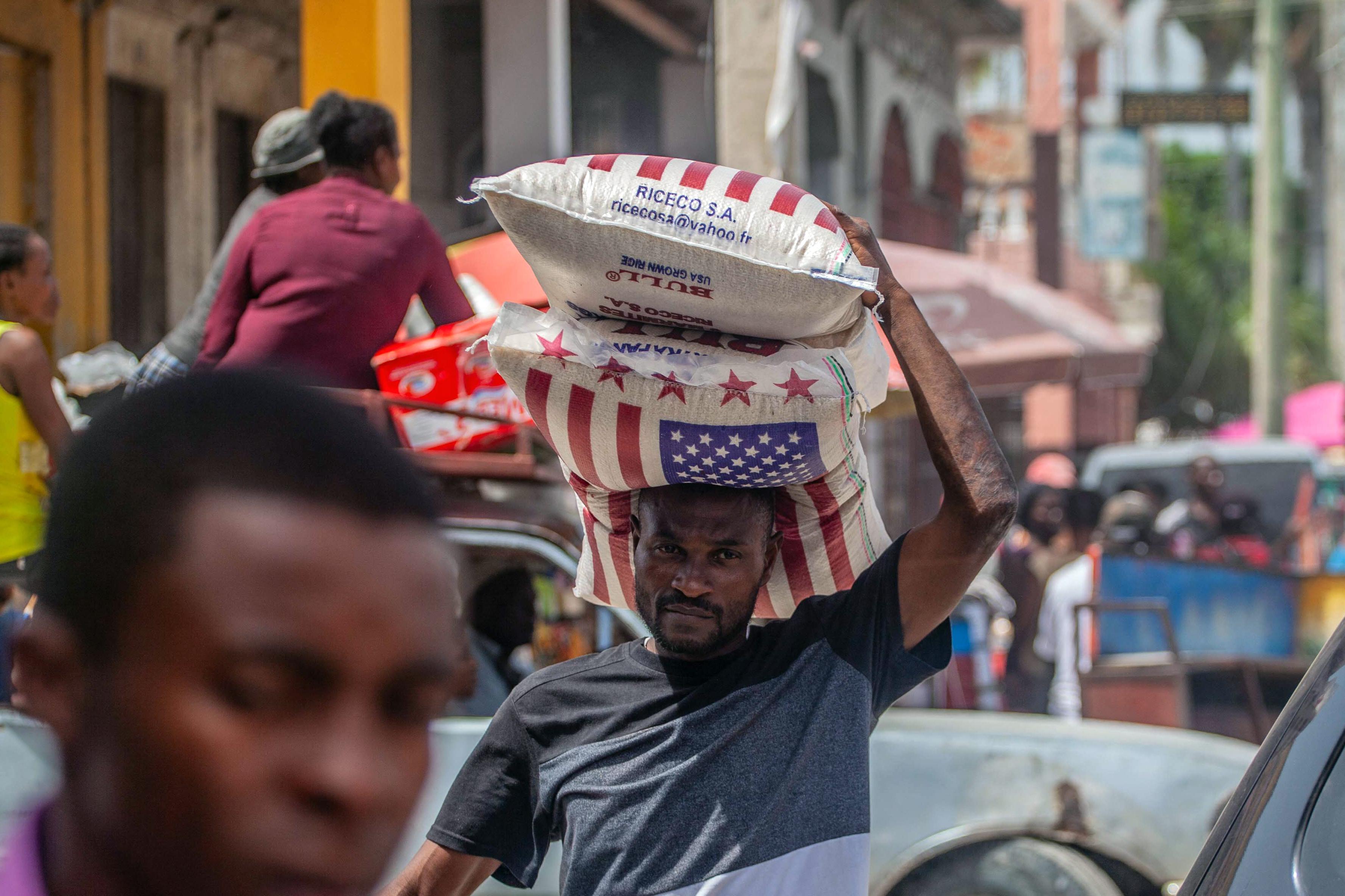 A man walks down the street carrying two bags on rice emblazoned with the American flag. 