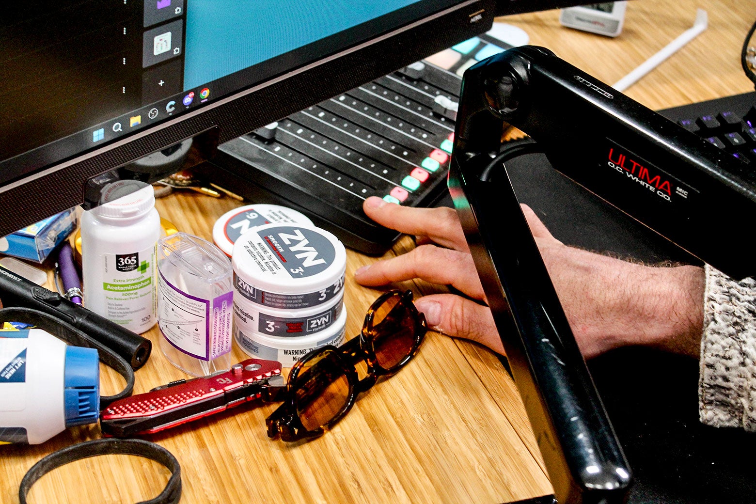 Items scattered on a desk, in front of a computer monitor, that include Zyn, a pair of sunglasses, bottles of vitamins and Advil, and a pocketknife.