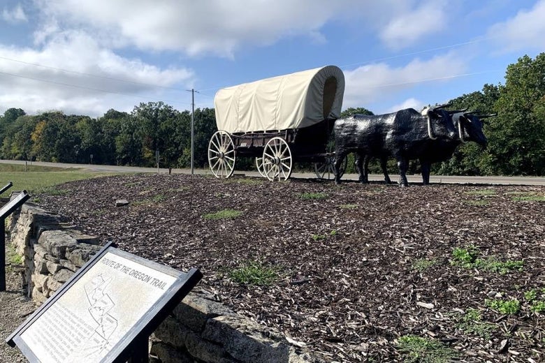 Two metal cattle pull a covered wagon in front of placards.