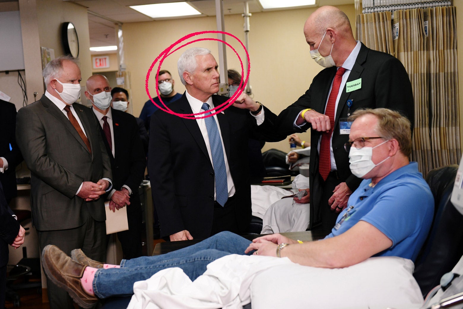 Mike Pence stands beside a masked patient in an exam room, surrounded by men wearing suits and surgical masks. Pence is the only person not wearing a mask.
