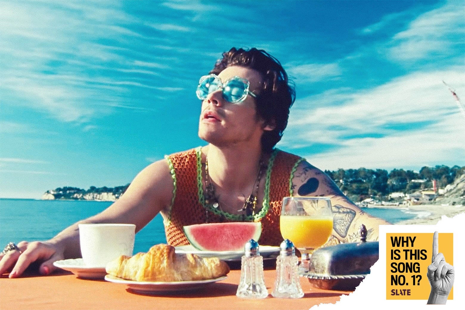 Still from the "Watermelon Sugar" video in which Harry Styles, wearing sunglasses, sits at a table on the beach with a slice of watermelon, a croissant, orange juice, a coffee cup, a butter dish, and salt and pepper shakers in front of him