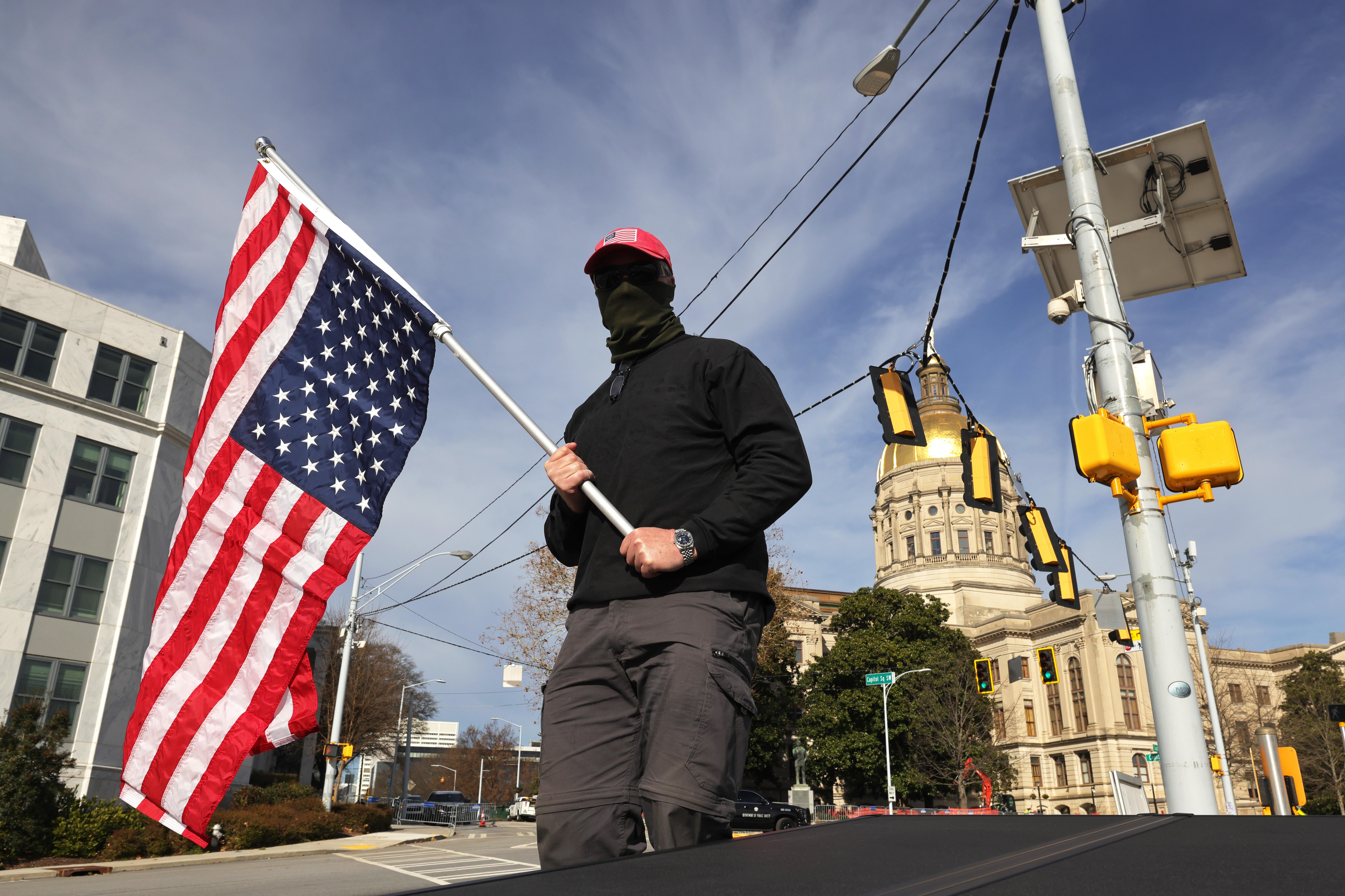 Man wearing black clothing, a black gaiter, and a red MAGA hat holds an upside-down American flag as he stands at an intersection. The Capitol can be seen in the distance behind him.