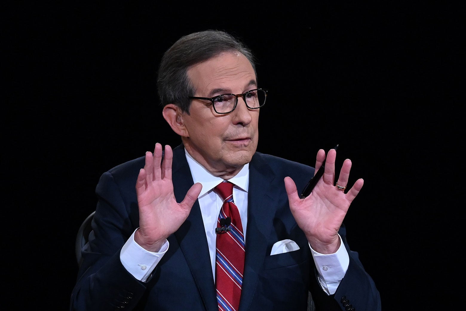 Chris Wallace, wearing black eyeglasses and a navy suit with a red tie, gestures with both hands palm-out, a pen tucked in his right fingers.