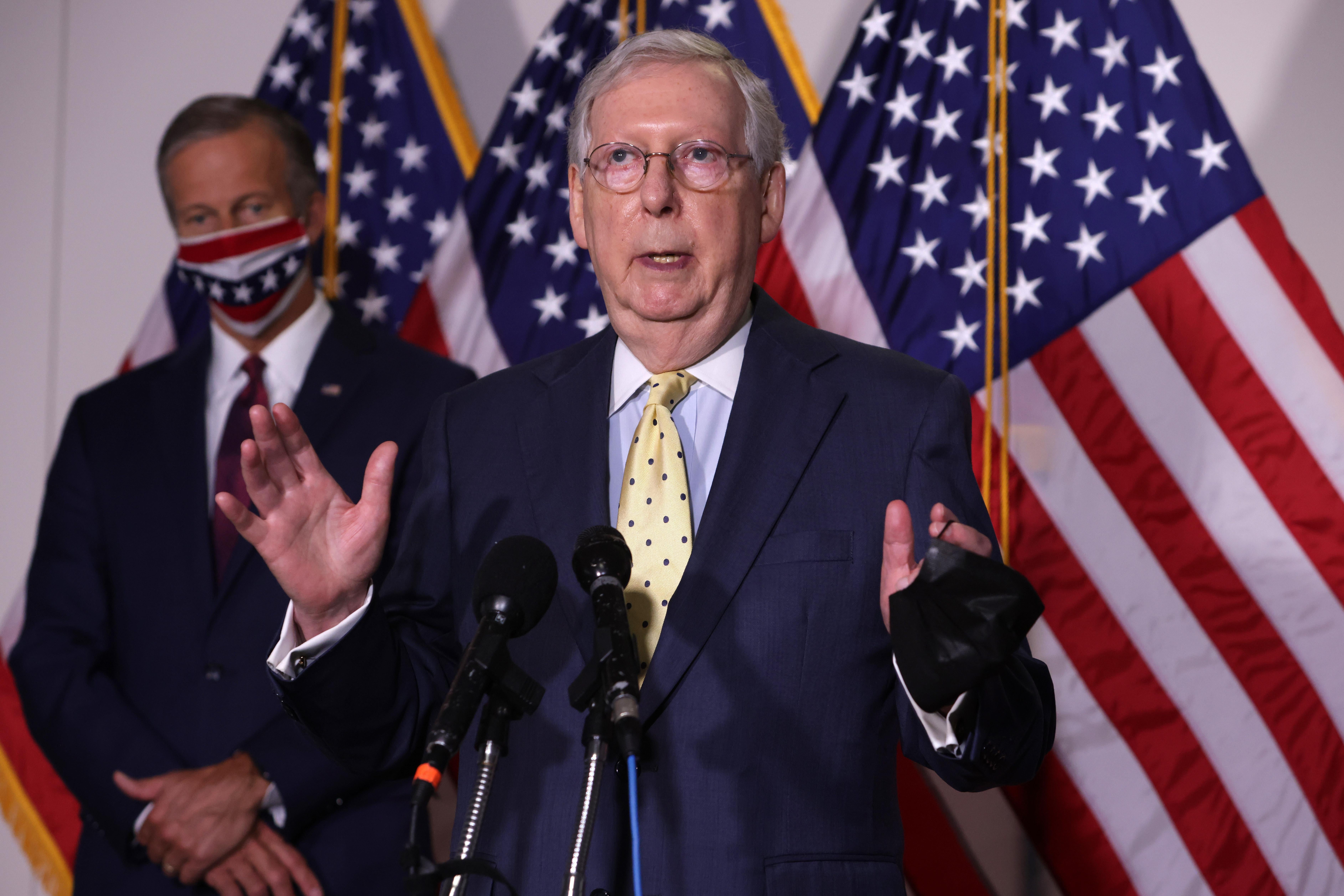 Mitch McConnell speaks into two microphones while standing in front of John Thune and some American flags.