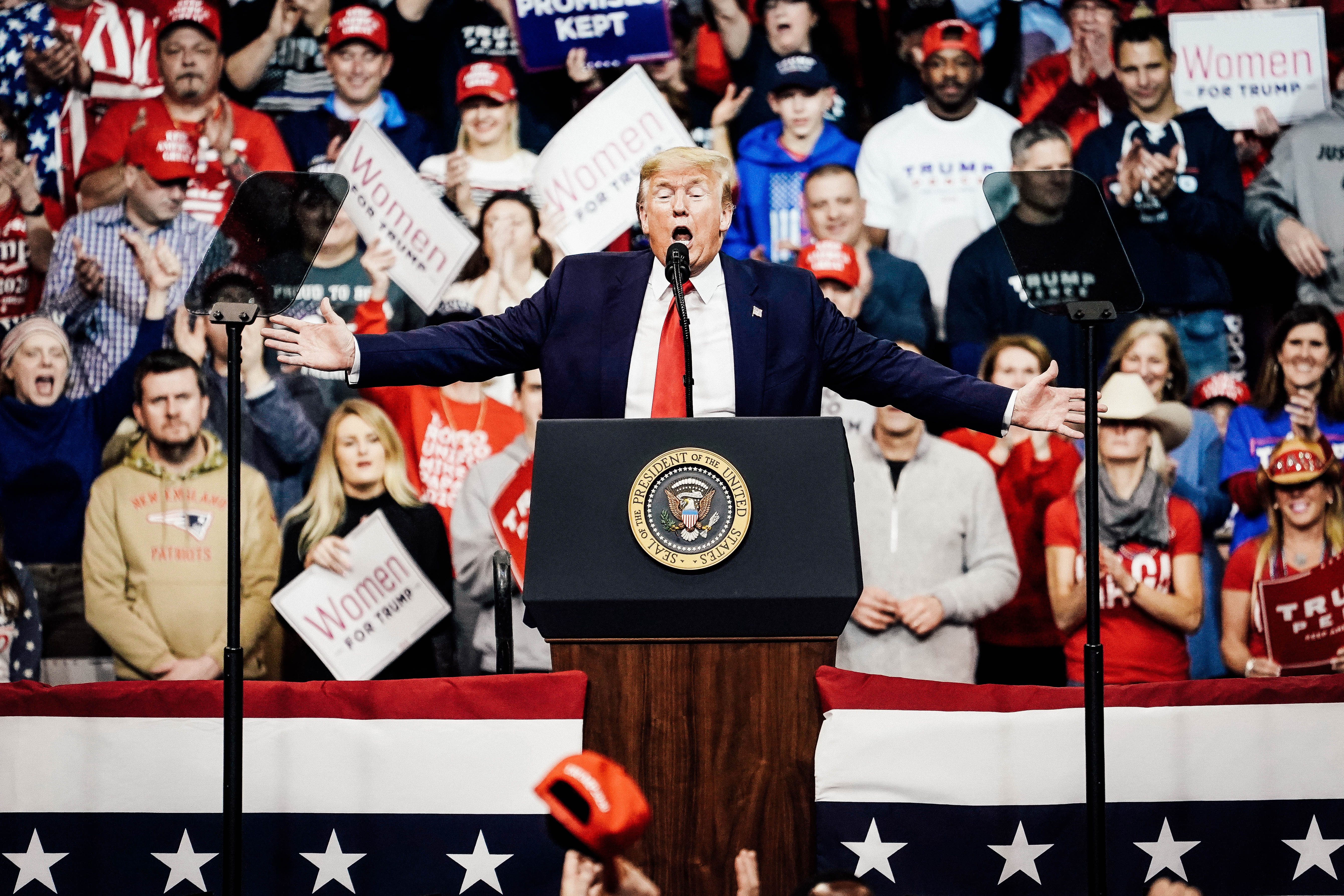 Trump stretches his hands while speaking at a lectern at a rally. A crowd is seen in the stands behind him.