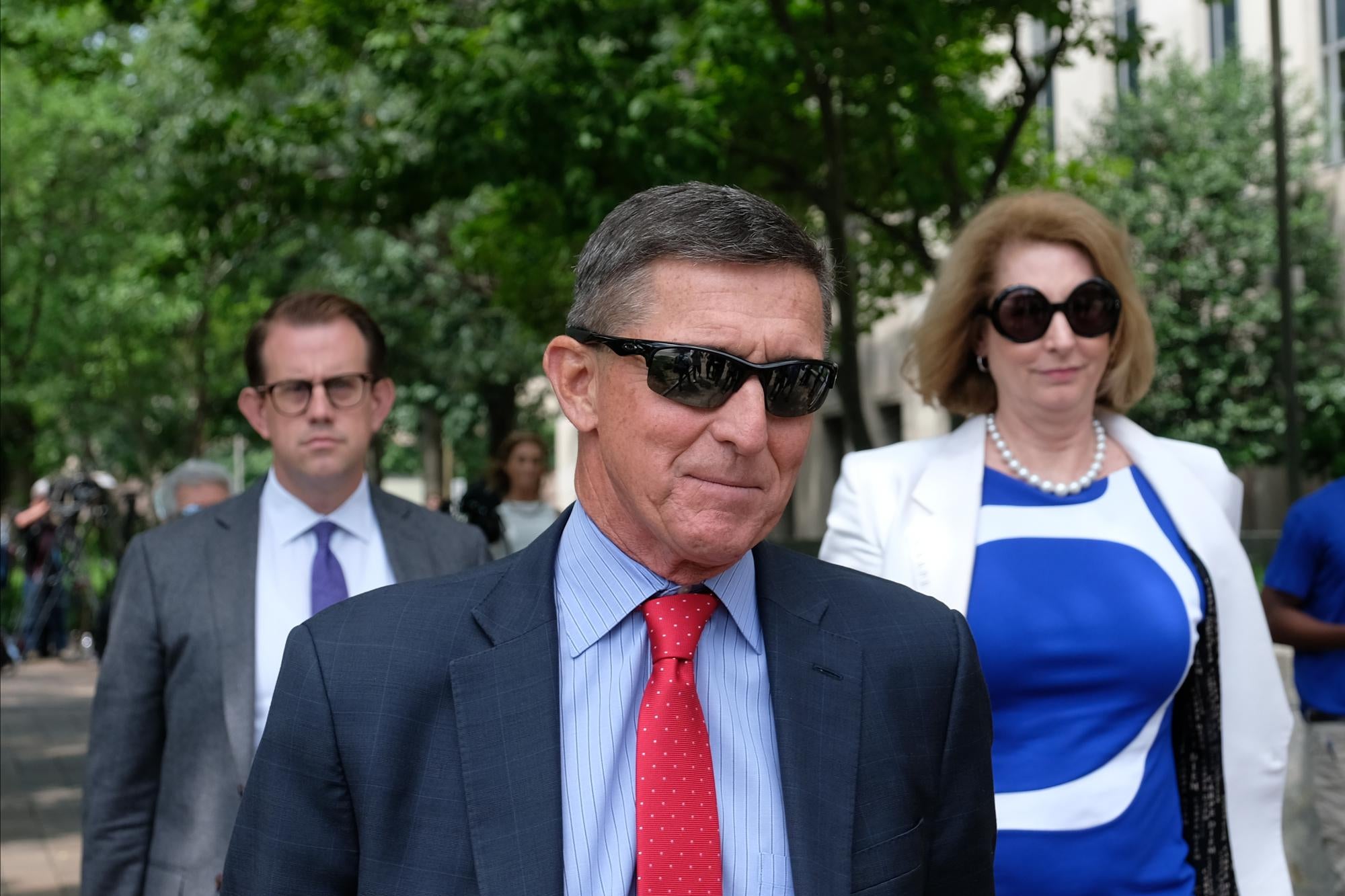 Michael Flynn, in sunglasses and a suit, walks with two people behind him.